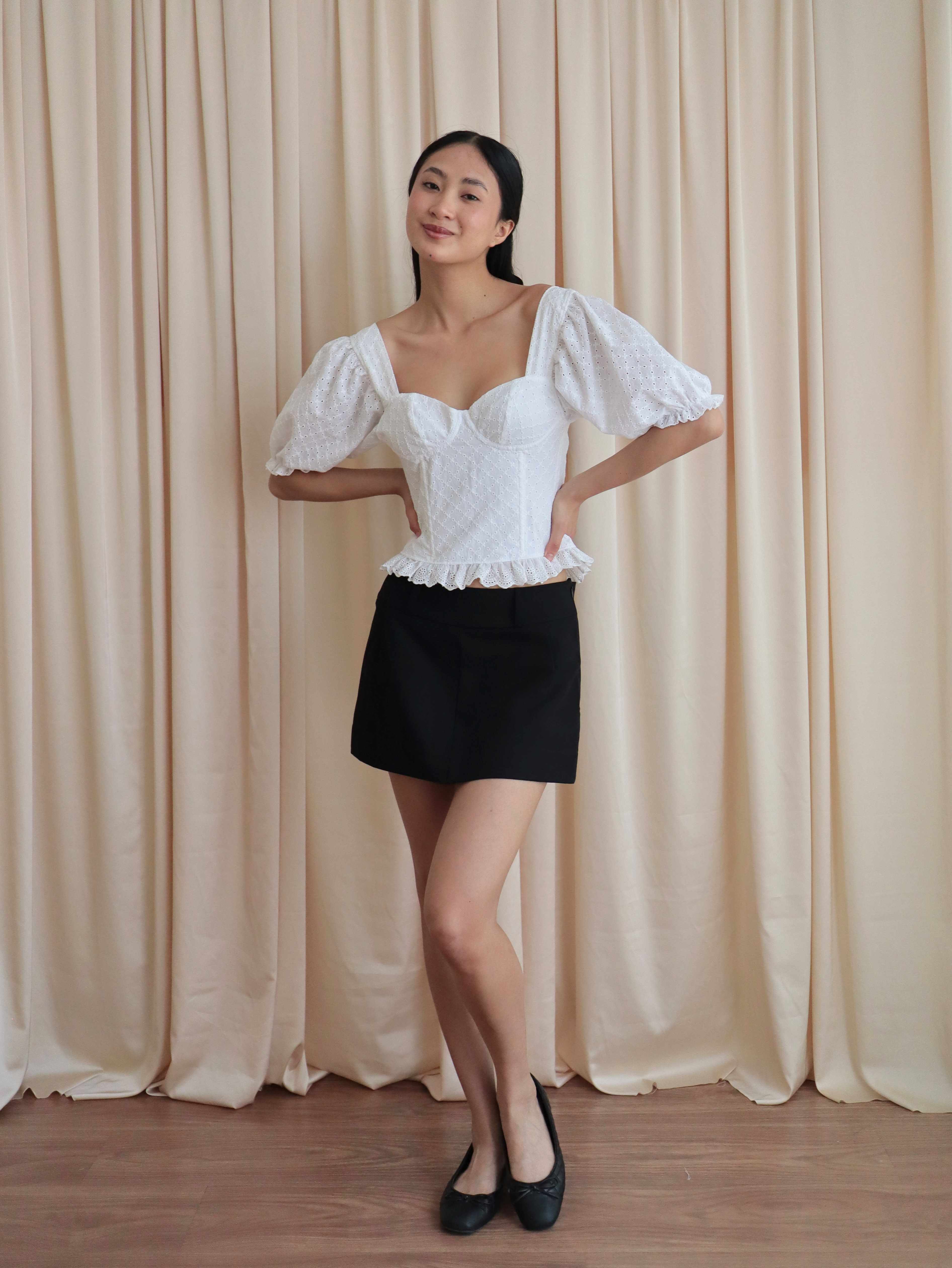 Mujer con corset broderie con escote corazón, mangas abullonadas y blonda usando falda negra corta con ballerinas. Posando en amarillo pastel.