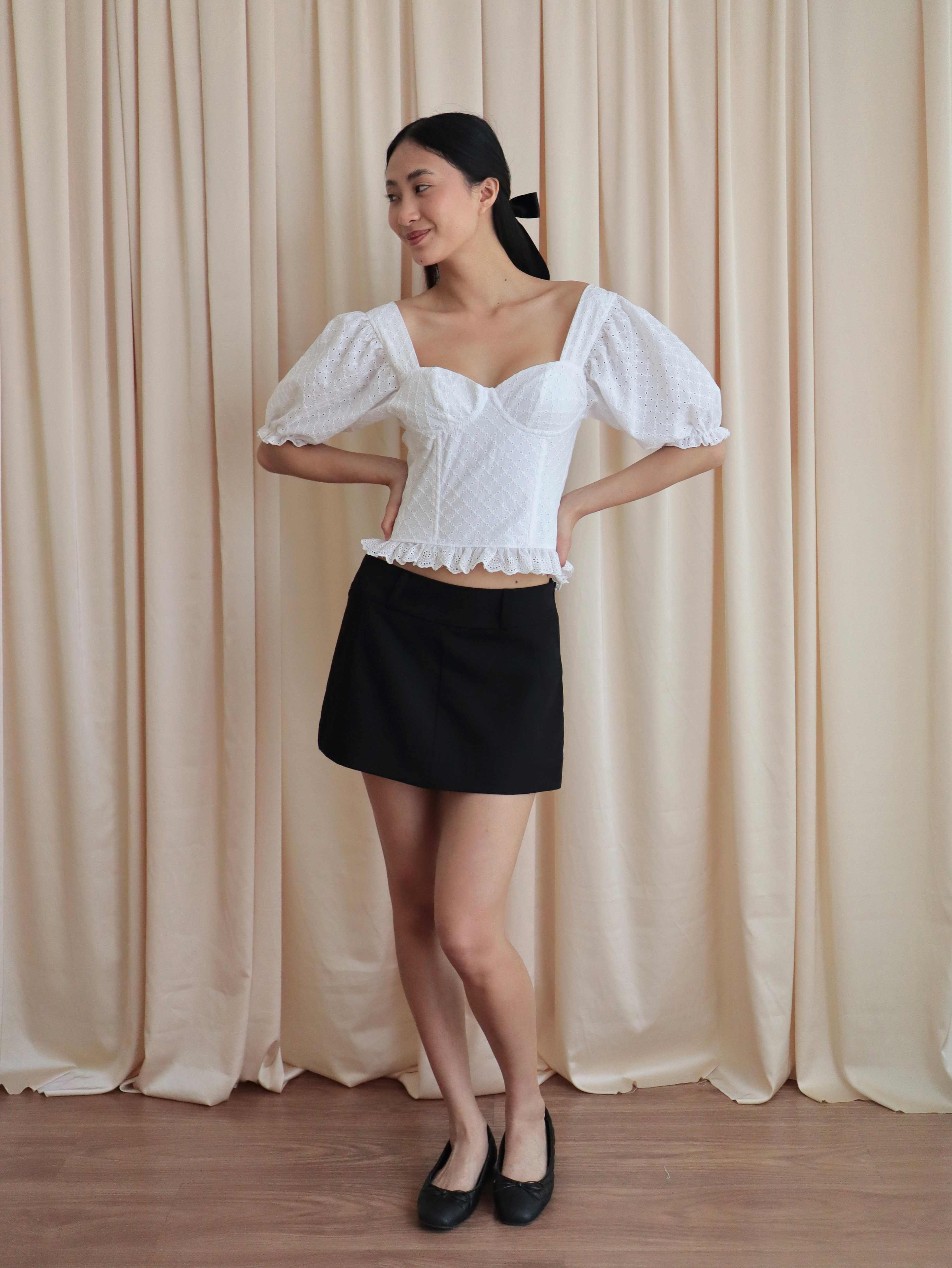 Mujer con corset broderie con escote corazón, mangas abullonadas y blonda usando falda negra corta con ballerinas. Posando en amarillo pastel.