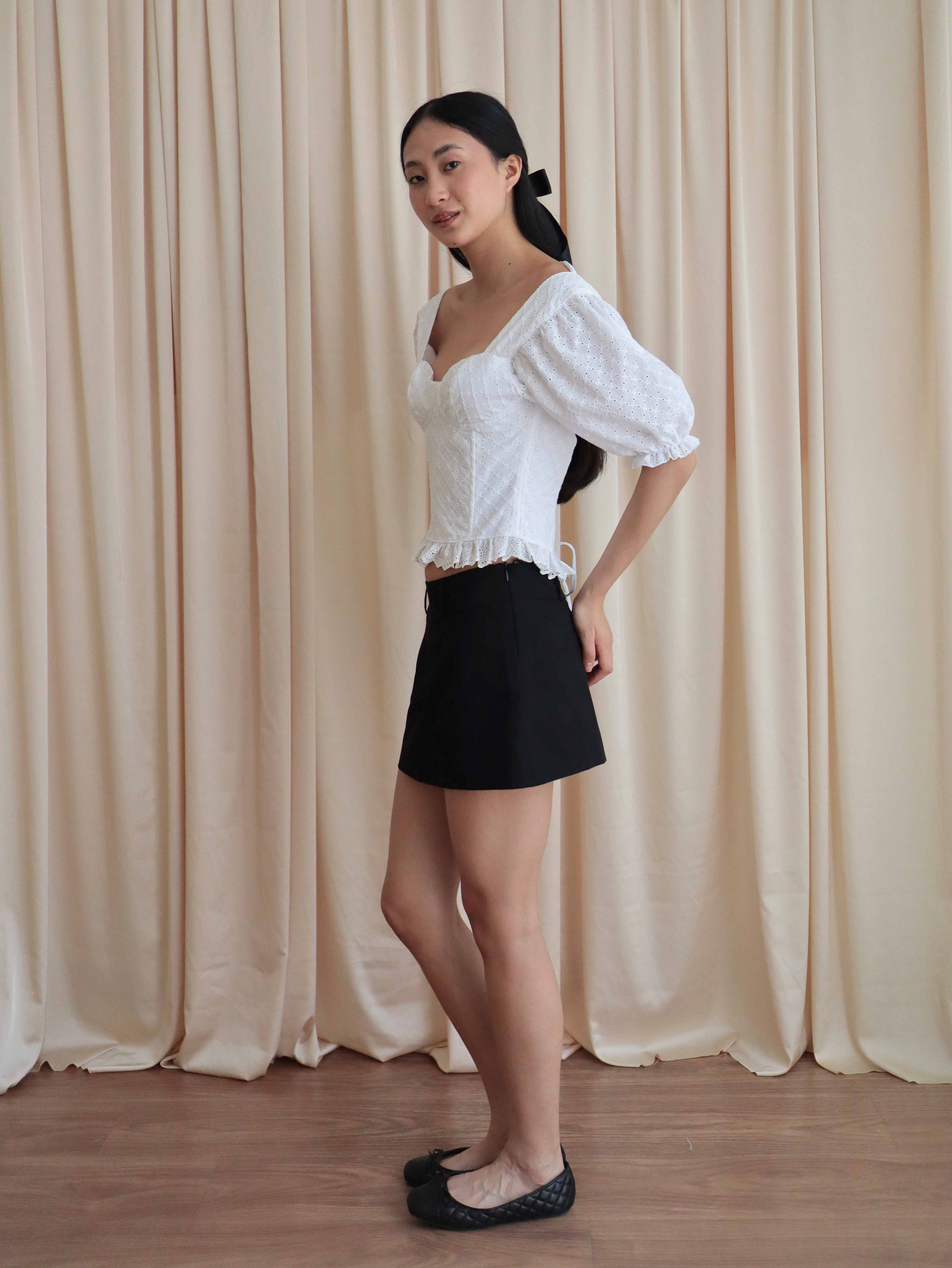 Mujer con corset broderie con escote corazón, mangas abullonadas y blonda usando falda negra corta con ballerinas. Posando en amarillo pastel.