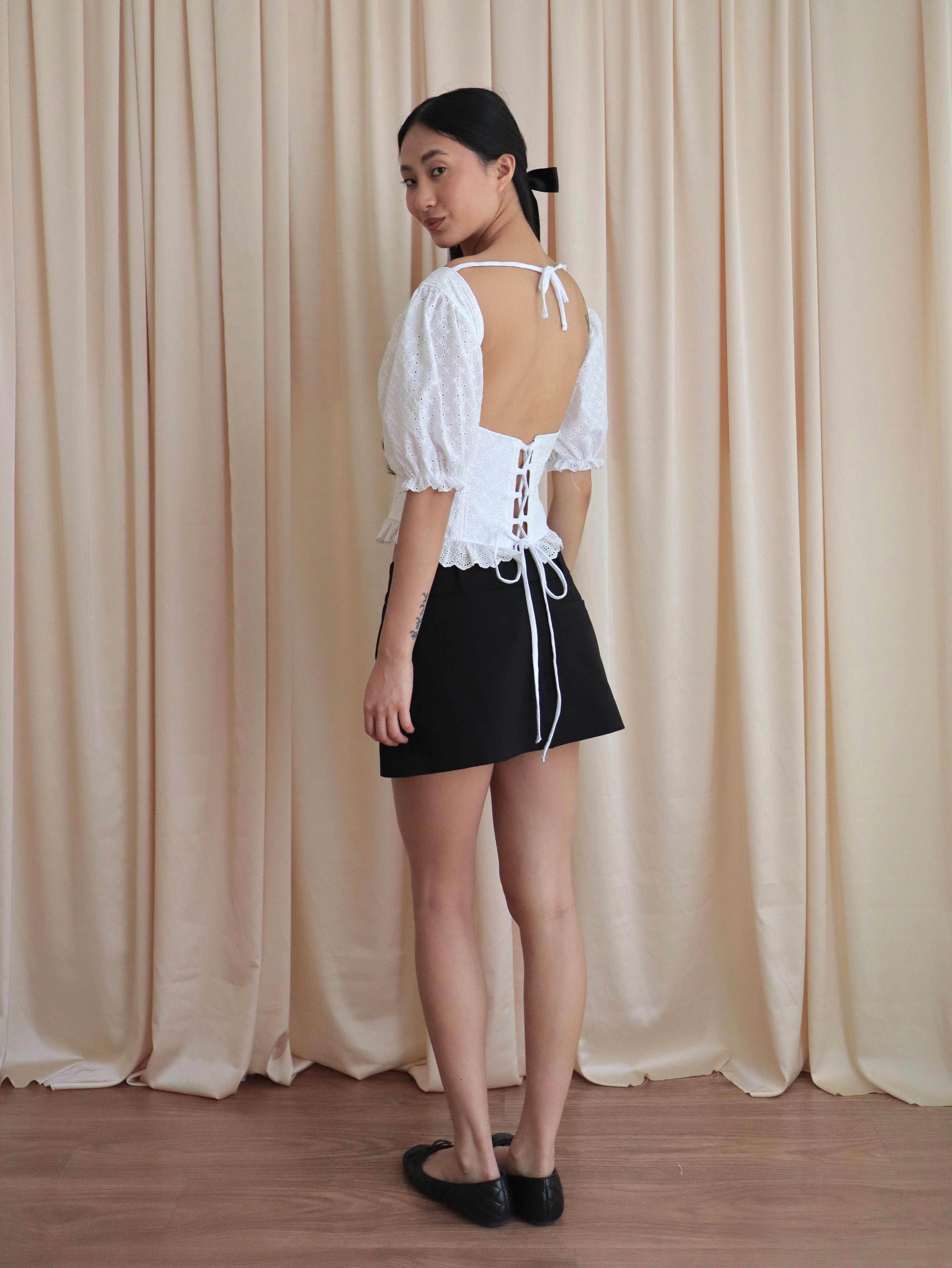 Mujer con corset broderie con escote corazón, mangas abullonadas y blonda usando falda negra corta con ballerinas. Posando en amarillo pastel.