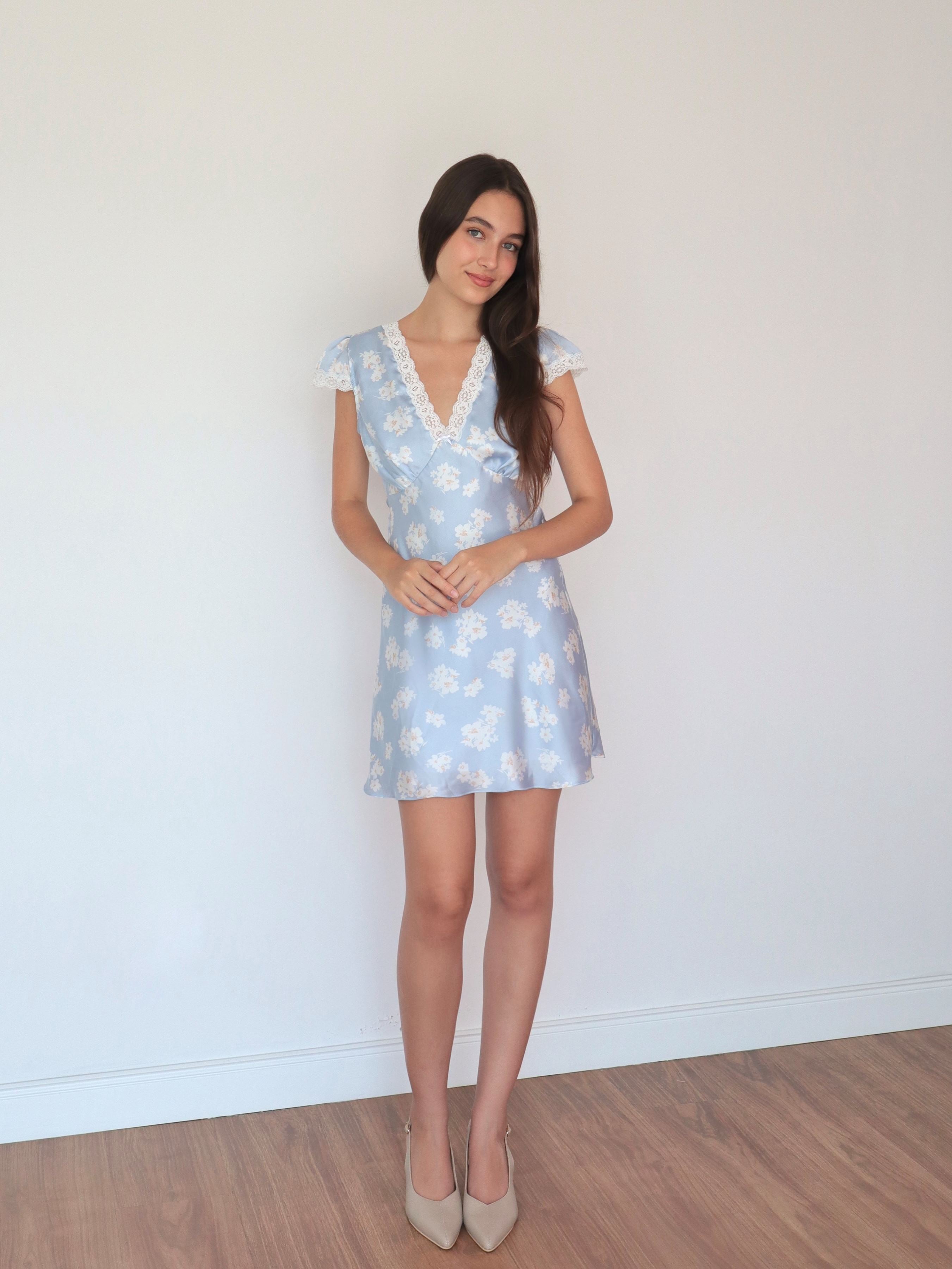 mujer en fondo blanco usando vestido escote V celeste, mangas cortas con blondas hecho de satin estampado con flores blancas estilo romántico