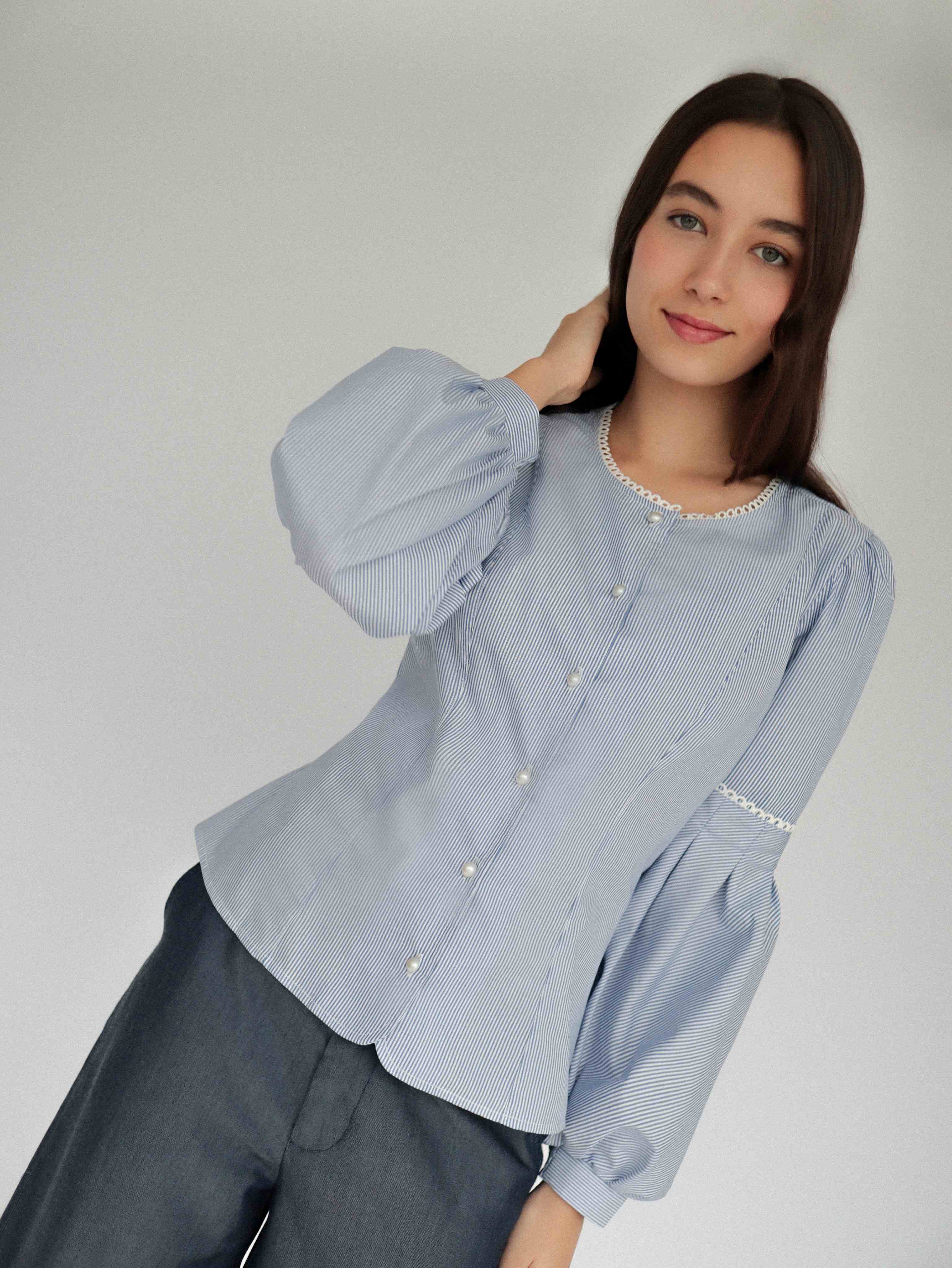 Mujer con blusa a rayas celeste con detalles tejidos, cuello redondo y mangas ligeramente voluminosas.  Posando en fondo blanco.