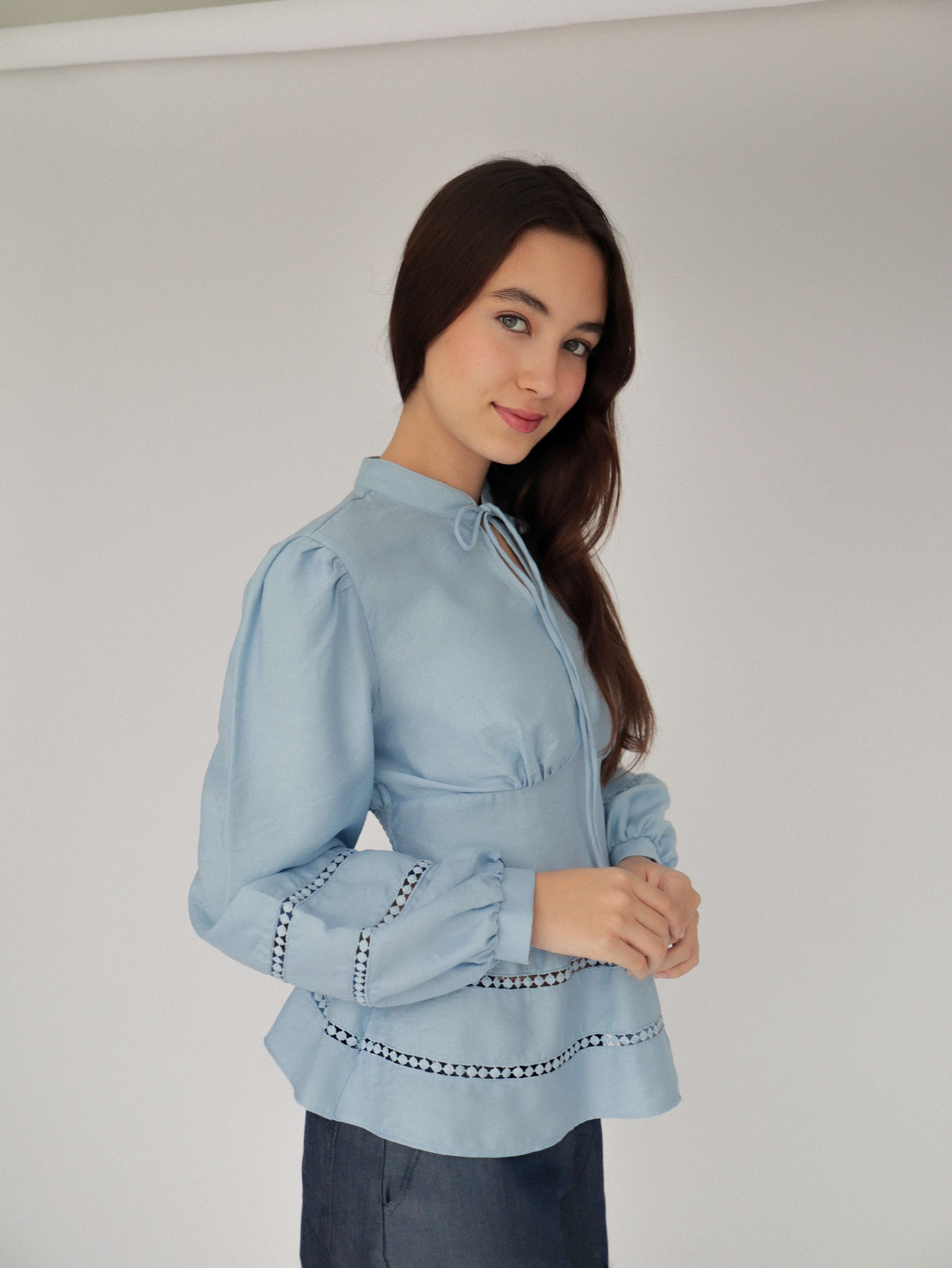 Mujer con blusa celeste con detalles tejidos y pantalones estilo denim posando en fondo blanco.
